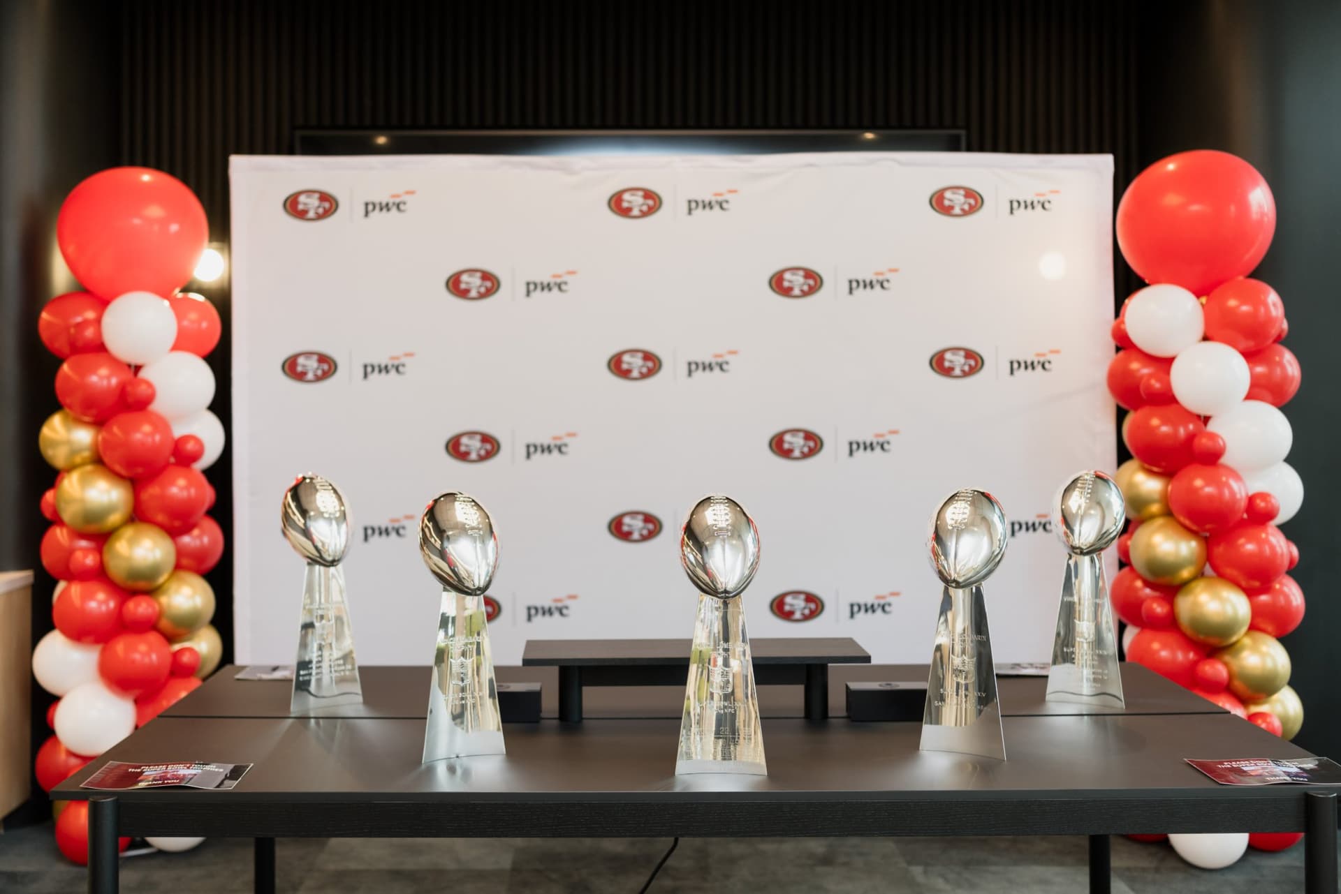 Five Super Bowl Lombardi Trophies displayed in front of the 49ers and PwC step-and-repeat with red, white and gold balloon columns at the PwC x 49ers event at PwC Silicon Valley in San Jose by JY Studio