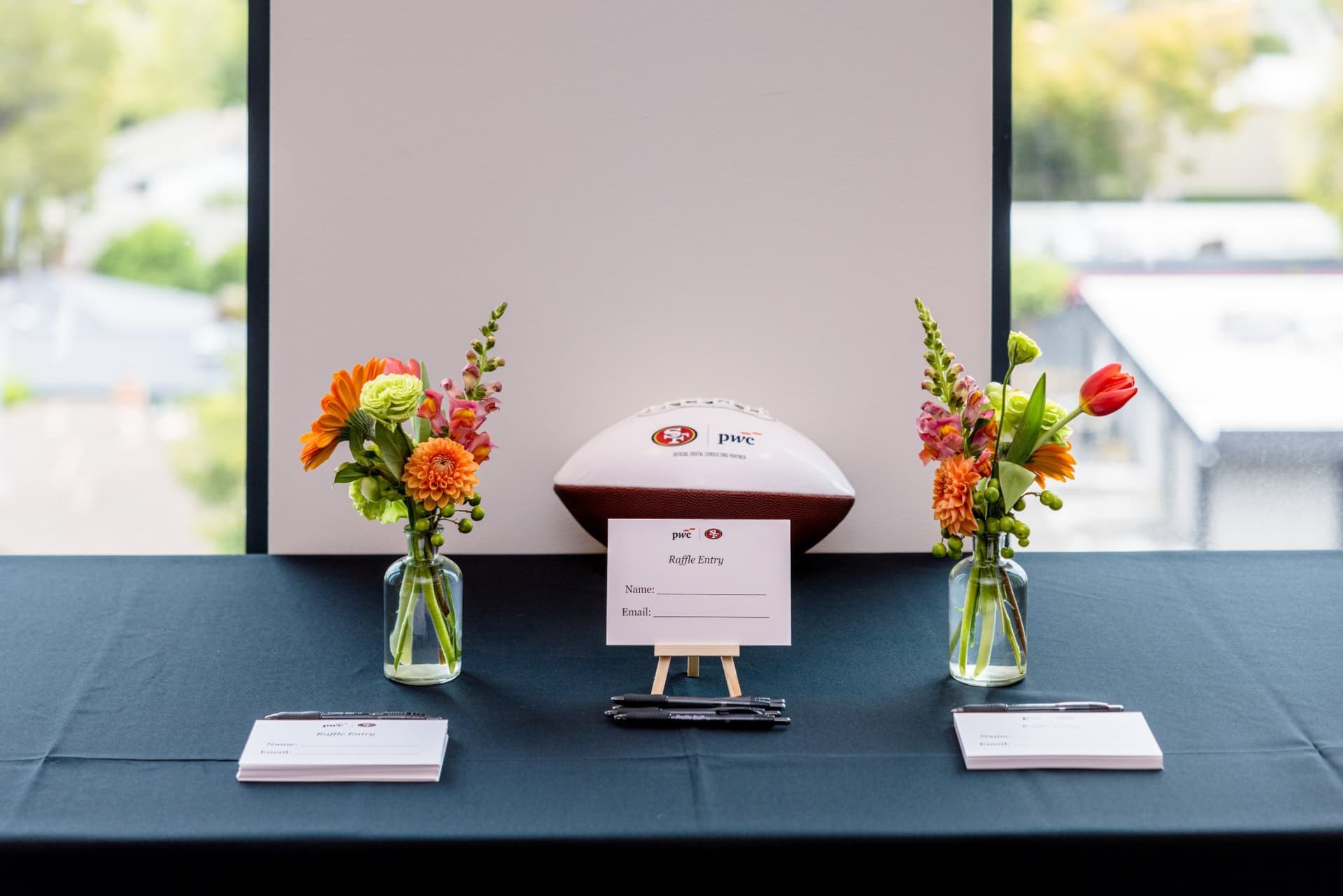 PwC-branded football and raffle entry cards on a table with fresh flower arrangements at the PwC x 49ers event at PwC Silicon Valley in San Jose by JY Studio