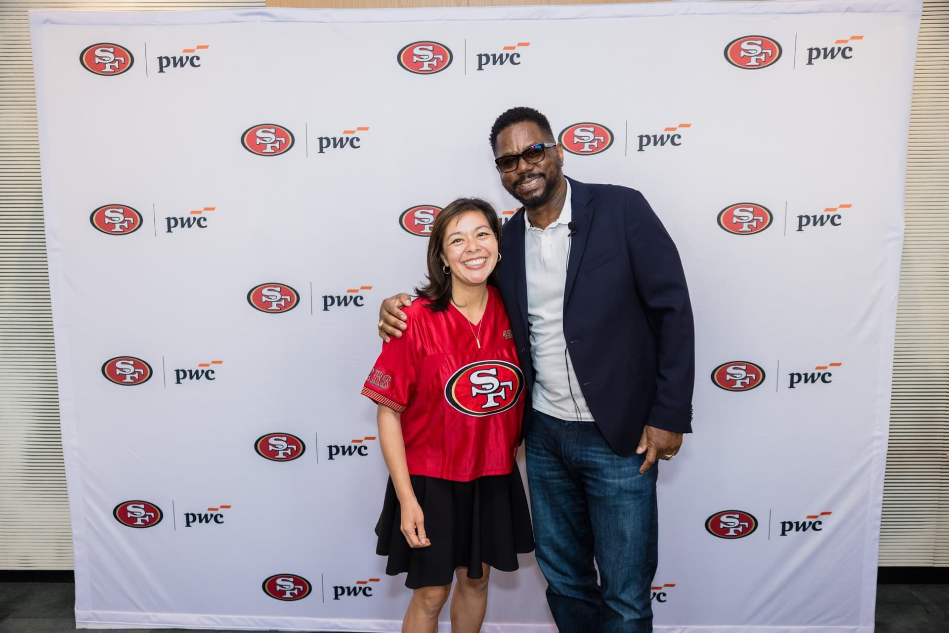 Fan in a red 49ers jersey posing with the 49ers legend at the step-and-repeat at the PwC x 49ers event at PwC Silicon Valley in San Jose by JY Studio