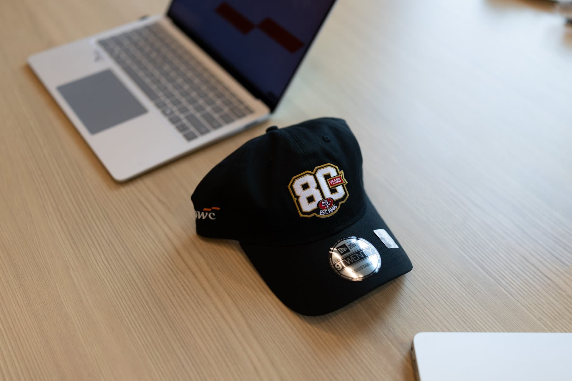 49ers 80th anniversary New Era cap on a desk beside a laptop at the PwC x 49ers event at PwC Silicon Valley in San Jose by JY Studio