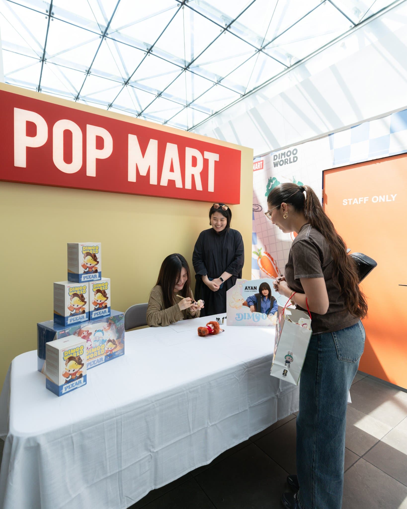 DIMOO artist Ayan signing for a fan under the red Pop Mart sign with DIMOO WORLD x PIXAR boxes on the table at the Pop Mart DIMOO signing event at Westfield Valley Fair in Santa Clara by JY Studio