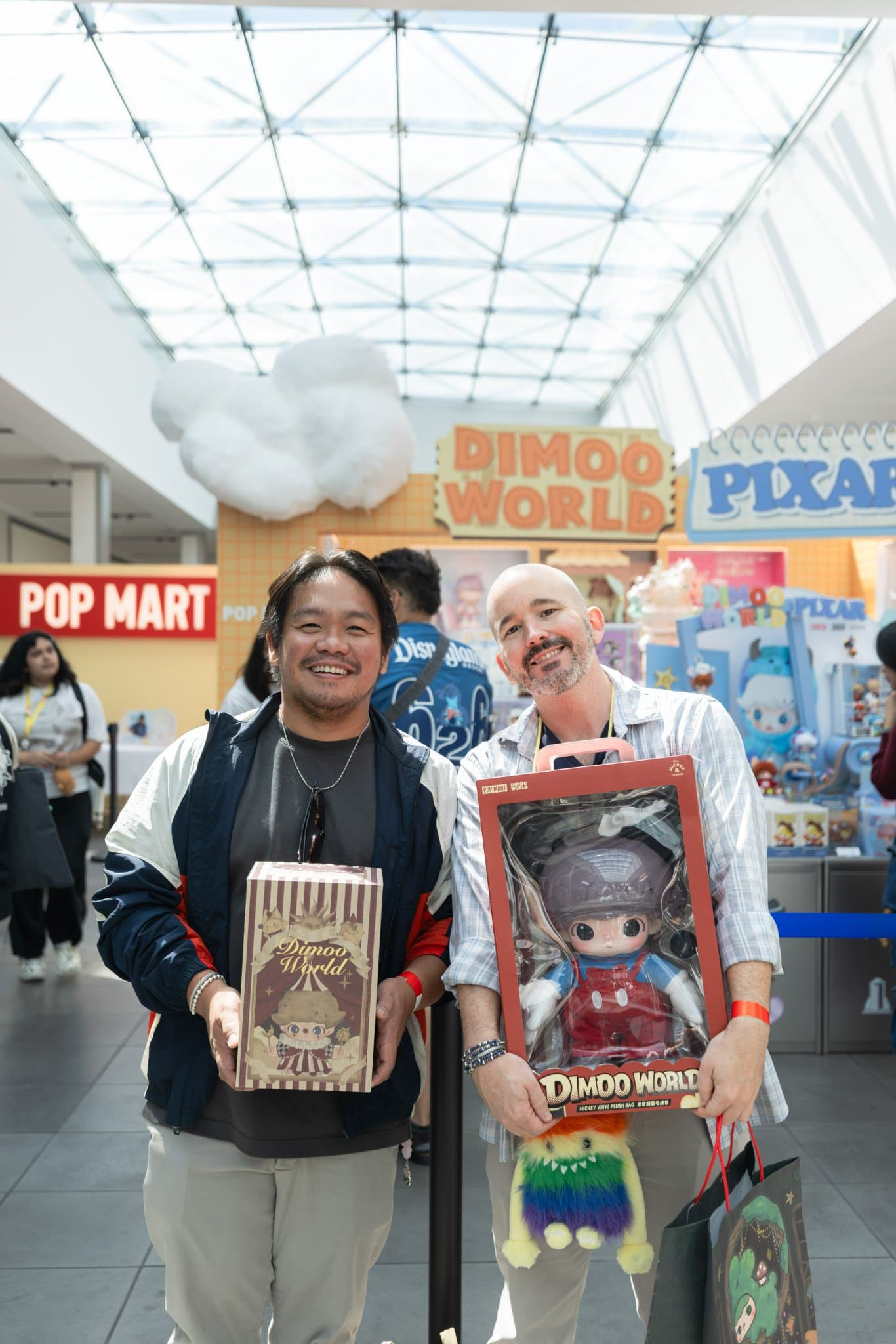 Two fans holding a boxed DIMOO WORLD doll and a DIMOO x PIXAR set in front of the pop-up at the Pop Mart DIMOO WORLD x PIXAR pop-up at Westfield Valley Fair in Santa Clara by JY Studio