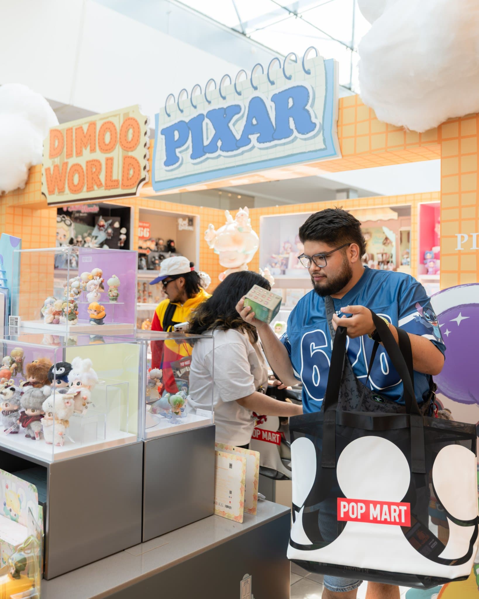 Fan carrying a Pop Mart tote bag in front of the DIMOO WORLD and PIXAR signs at the Pop Mart DIMOO WORLD x PIXAR pop-up at Westfield Valley Fair in Santa Clara by JY Studio