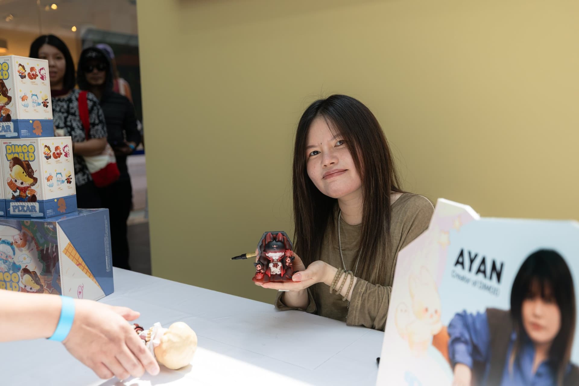 DIMOO creator Ayan smiling with a DIMOO figure beside the Ayan name card at the Pop Mart DIMOO signing event at Westfield Valley Fair in Santa Clara by JY Studio
