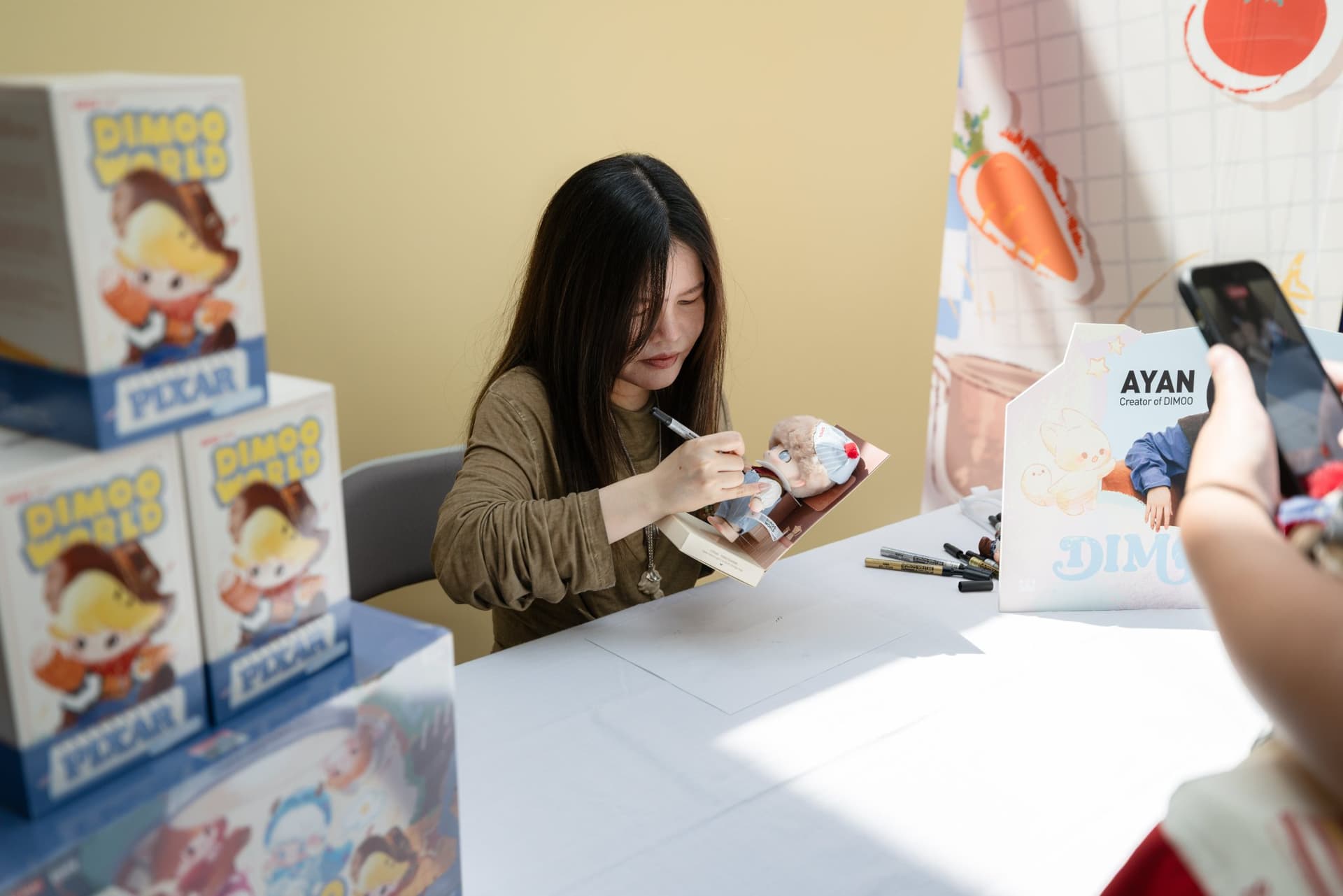 DIMOO creator Ayan signing a DIMOO x PIXAR figure for a collector at the Pop Mart DIMOO signing event at Westfield Valley Fair in Santa Clara by JY Studio
