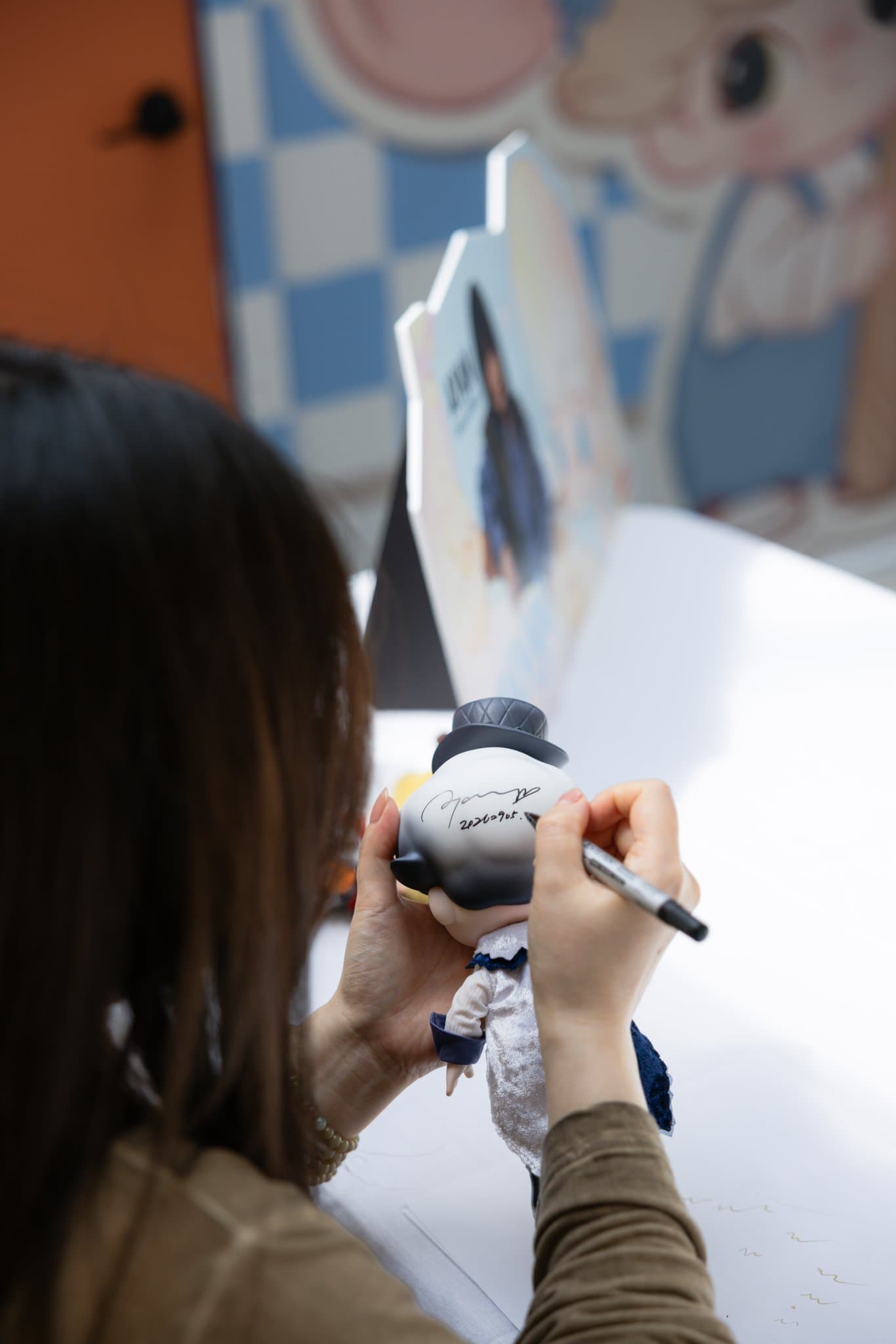 Close-up of DIMOO artist Ayan hand-signing a DIMOO figure with a marker at the Pop Mart DIMOO signing event at Westfield Valley Fair in Santa Clara by JY Studio