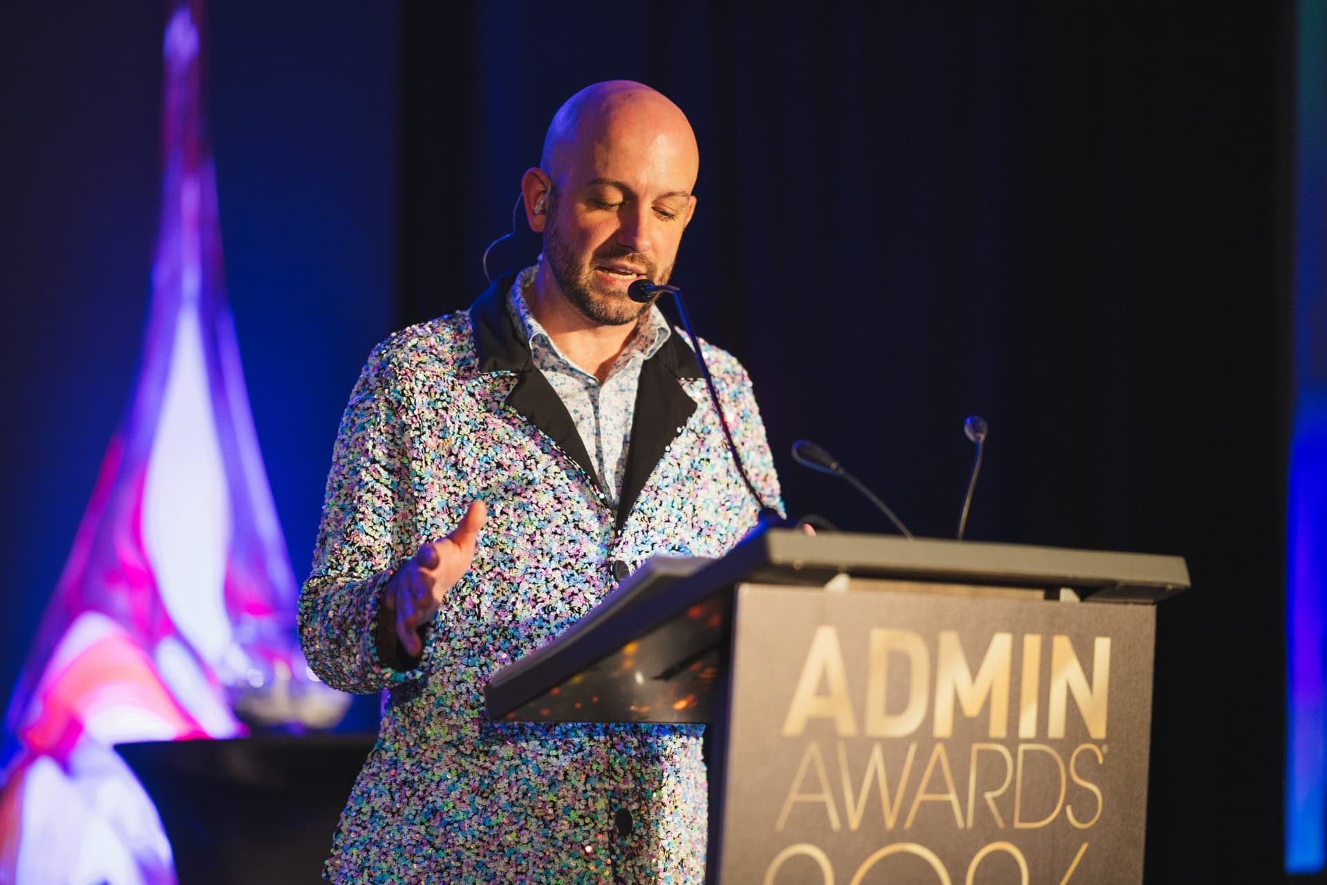 Emcee in a sequined suit reading from the Admin Awards 2026 podium during the awards ceremony at Admin Awards 2026 at SFO Marriott Waterfront, Burlingame by JY Studio
