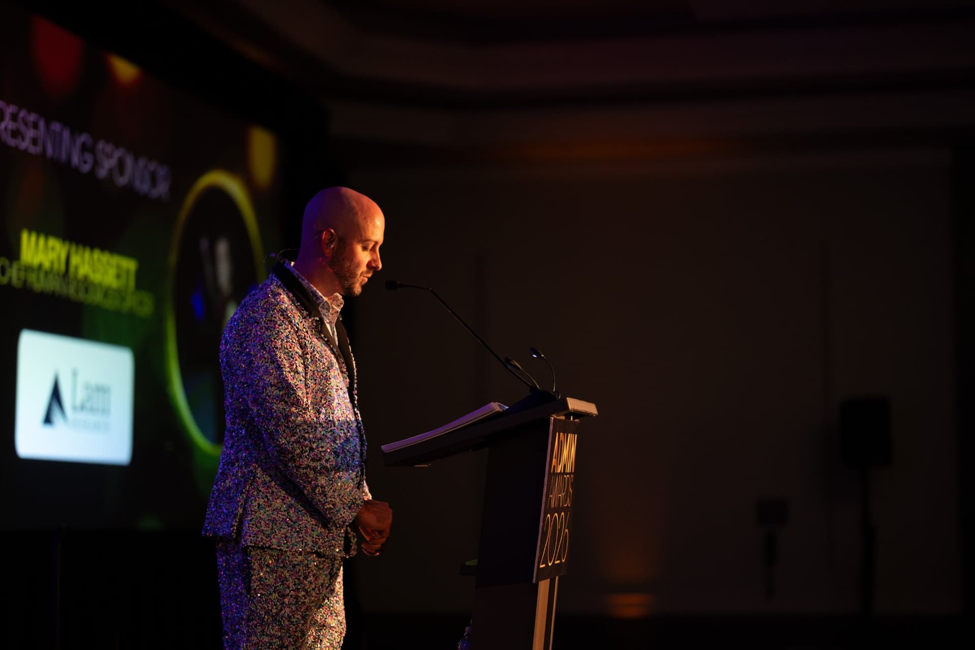 Emcee at the lectern beside the presenting sponsor screen at Admin Awards 2026 at SFO Marriott Waterfront, Burlingame by JY Studio