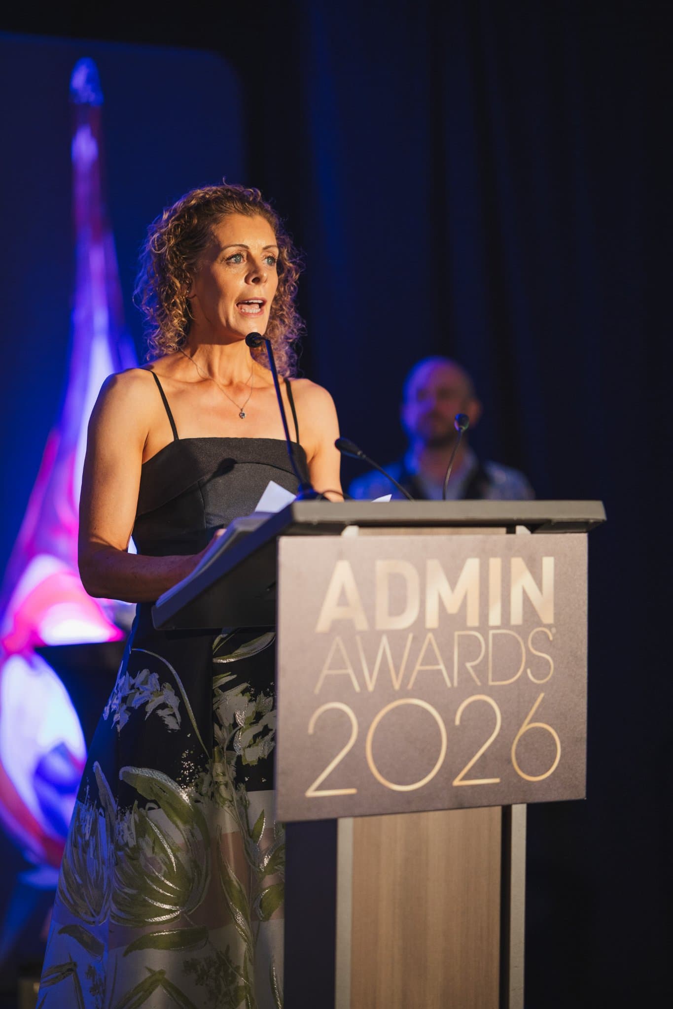 Speaker in a black floral gown addressing the gala audience from the podium at Admin Awards 2026 at SFO Marriott Waterfront, Burlingame by JY Studio