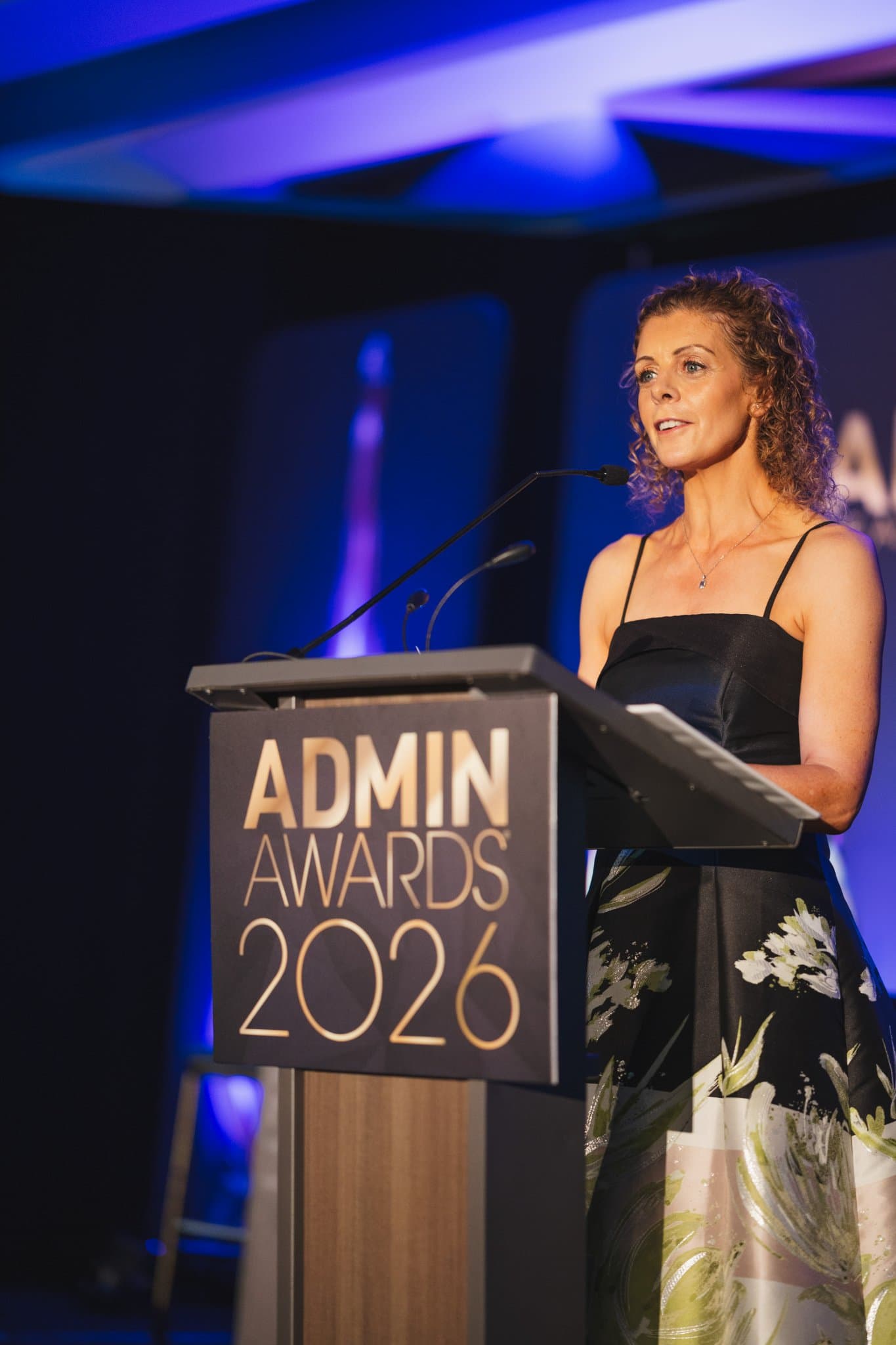 Close-up of a speaker at the Admin Awards 2026 podium under blue stage lighting at Admin Awards 2026 at SFO Marriott Waterfront, Burlingame by JY Studio