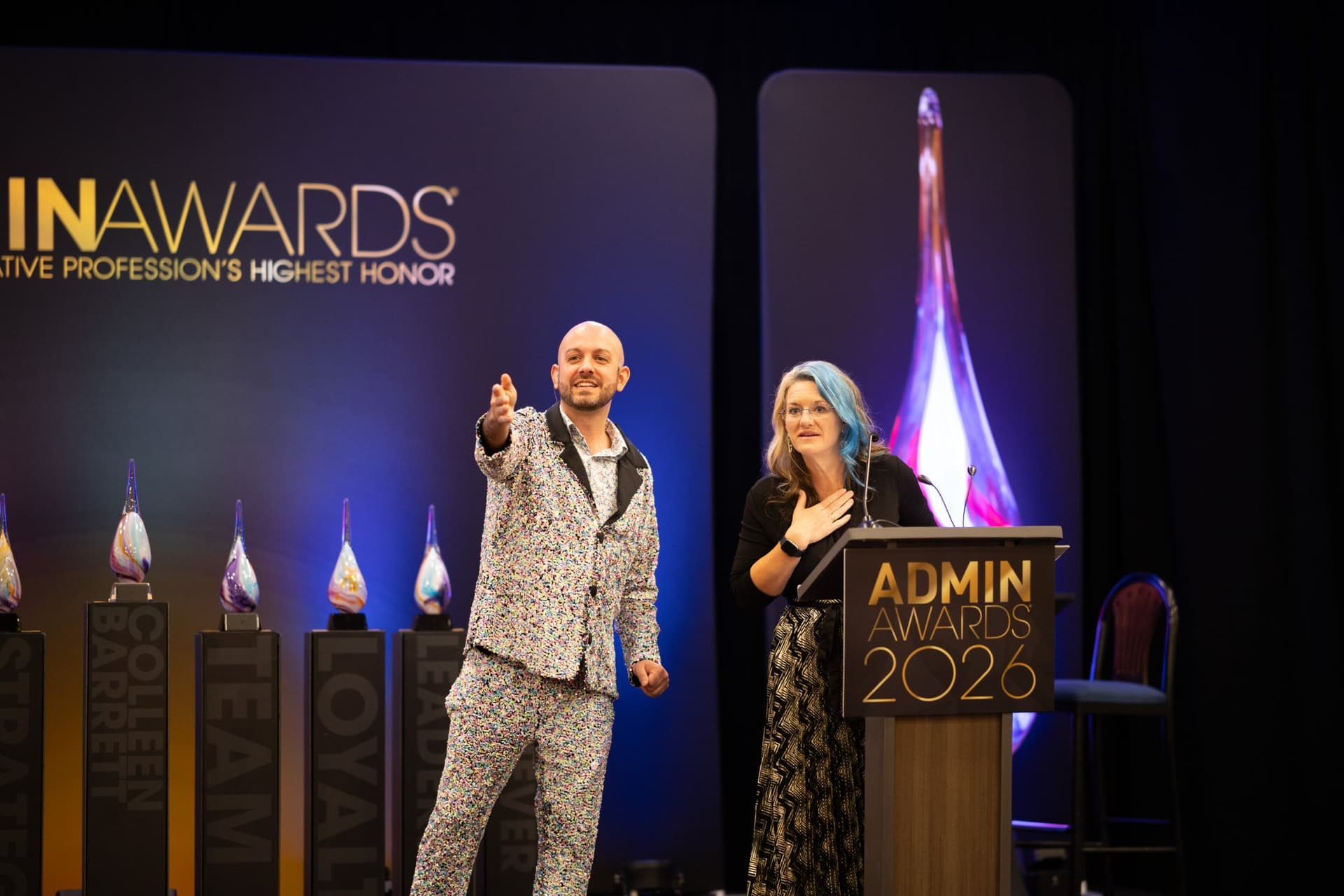 Emcee celebrating an honoree beside the podium in front of the award trophies at Admin Awards 2026 at SFO Marriott Waterfront, Burlingame by JY Studio