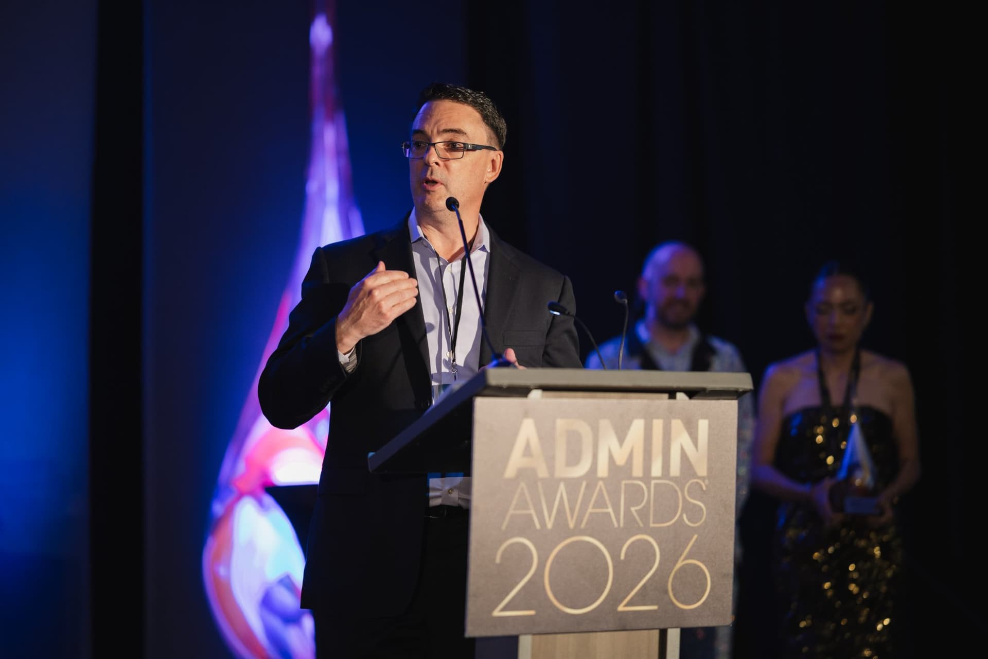 Presenter in a suit speaking at the podium as the honoree waits on stage at Admin Awards 2026 at SFO Marriott Waterfront, Burlingame by JY Studio