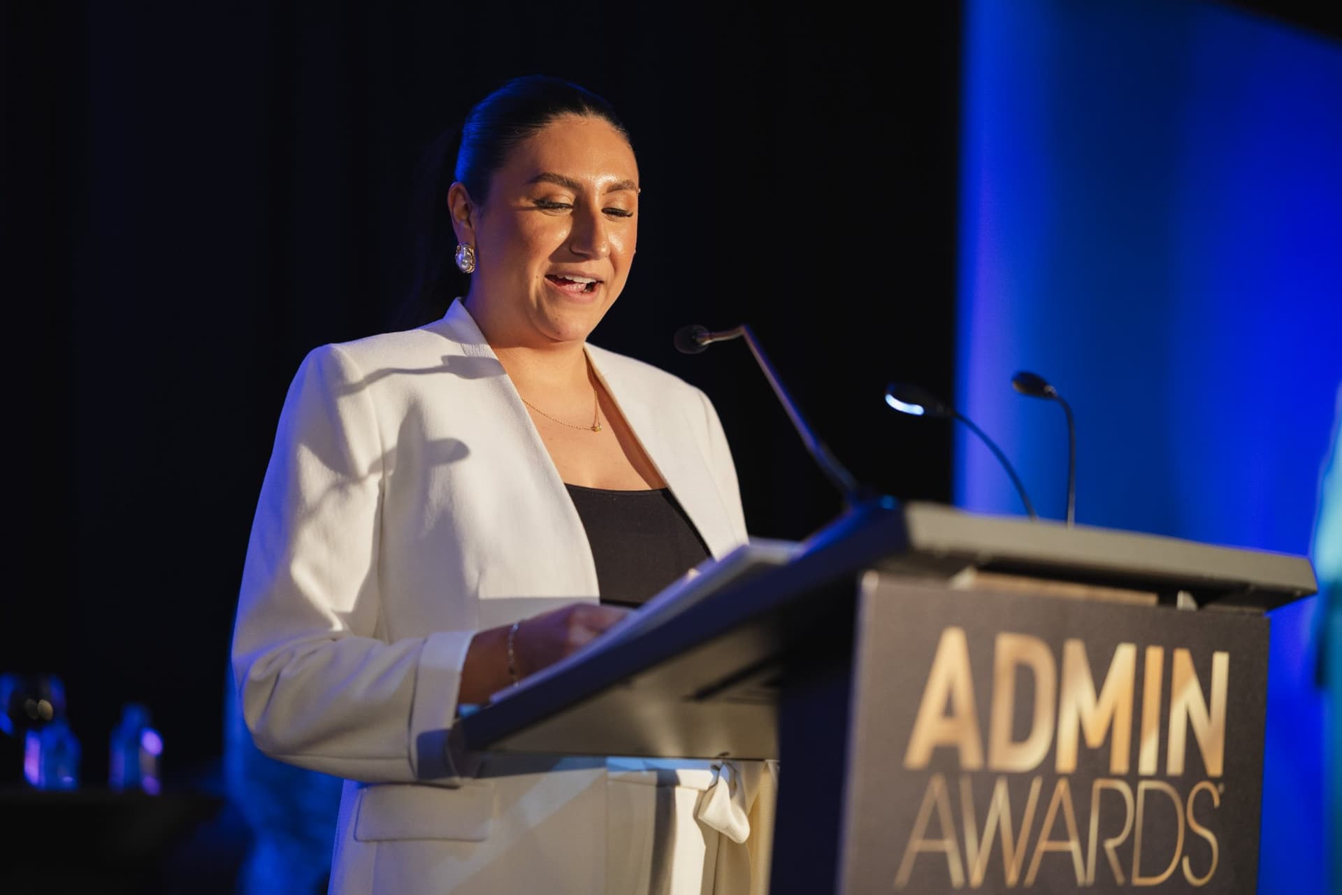 Presenter in a white blazer speaking at the Admin Awards podium at Admin Awards 2026 at SFO Marriott Waterfront, Burlingame by JY Studio