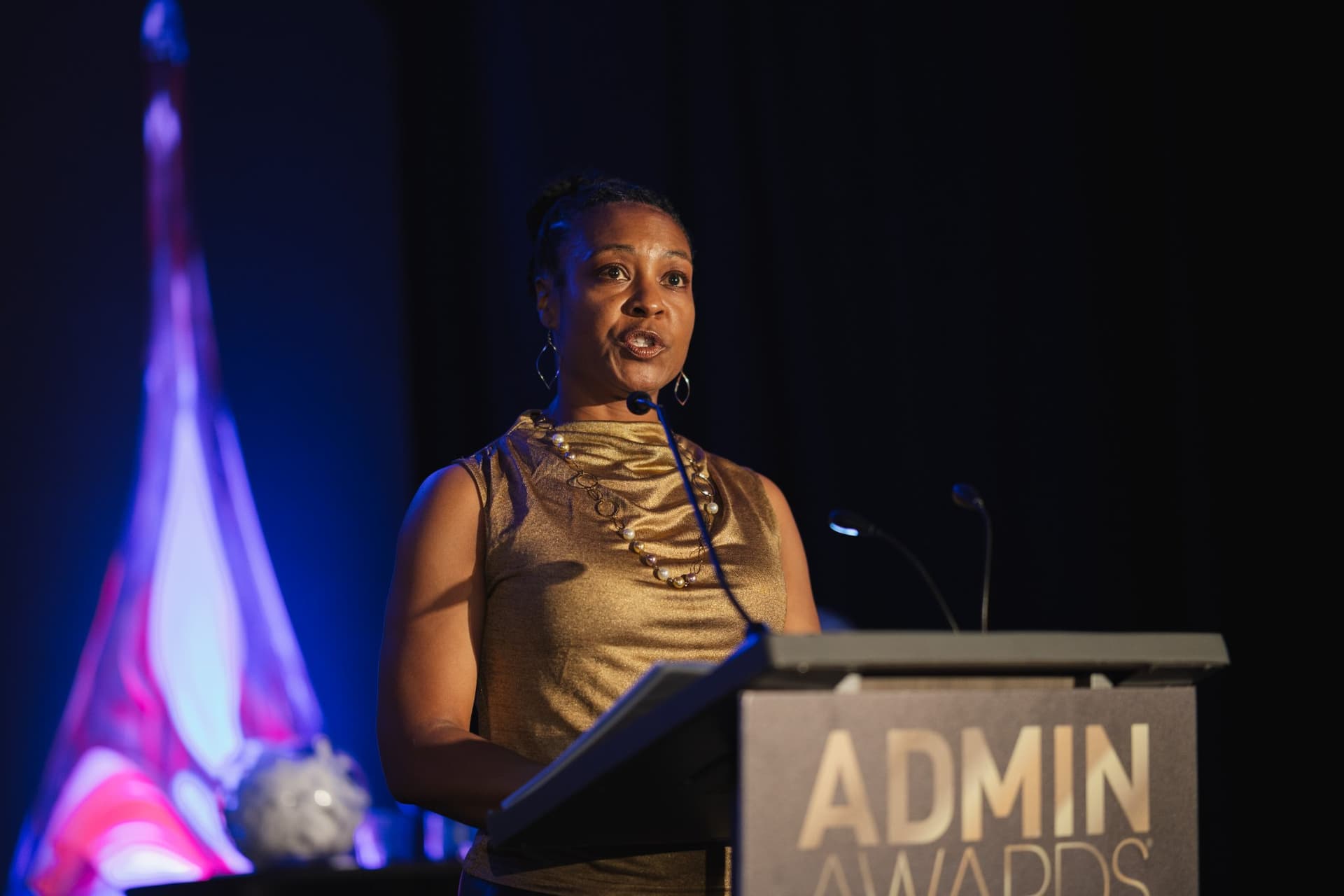 Speaker in a gold top addressing the audience from the Admin Awards podium at Admin Awards 2026 at SFO Marriott Waterfront, Burlingame by JY Studio
