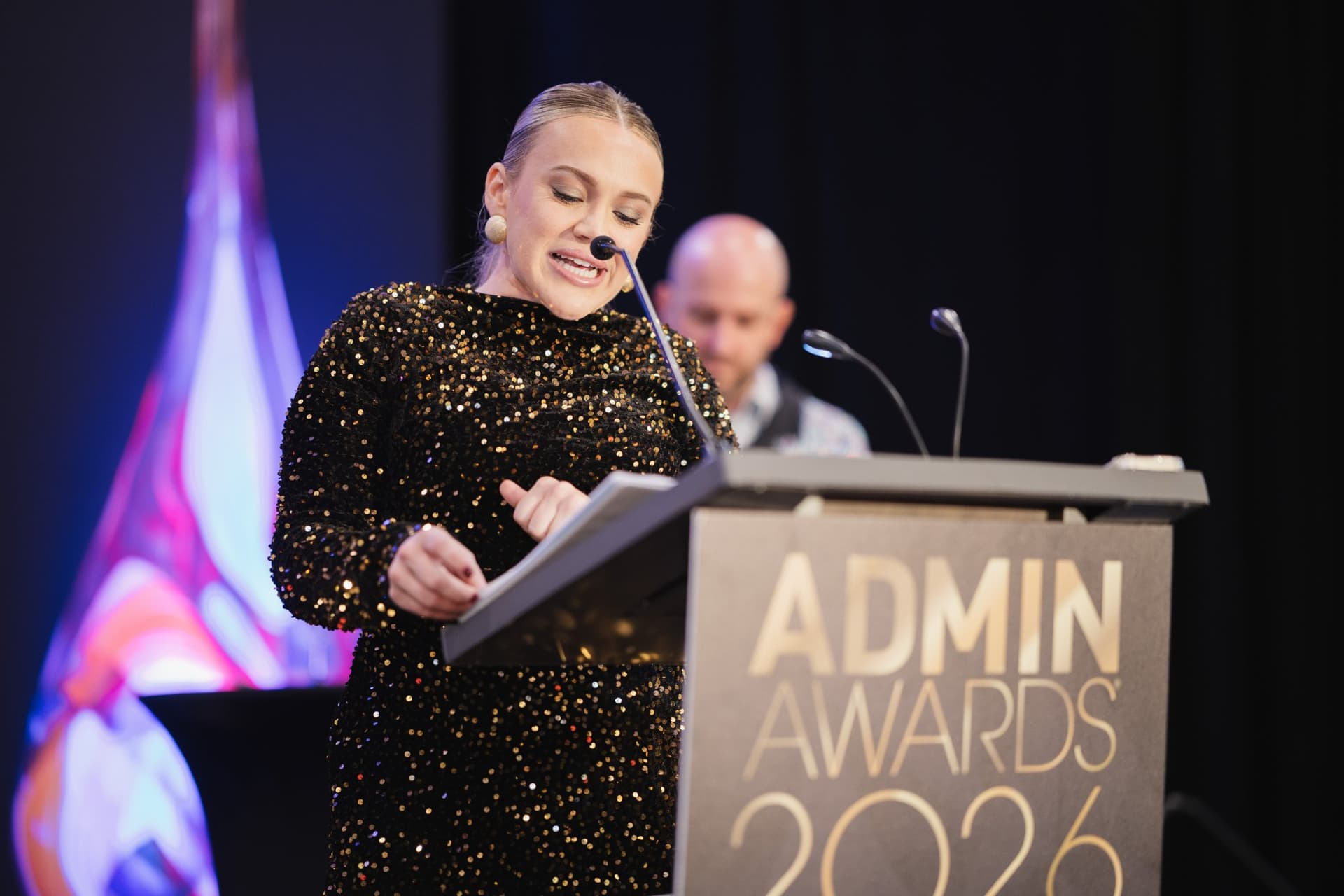 Speaker in a sequined dress reading from the Admin Awards 2026 podium at Admin Awards 2026 at SFO Marriott Waterfront, Burlingame by JY Studio