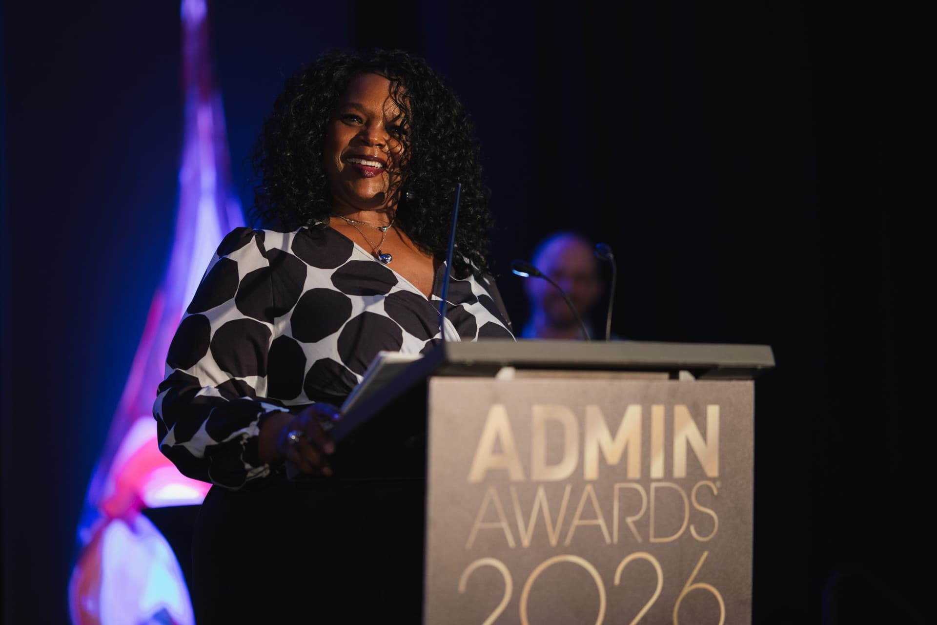 Smiling speaker in a polka-dot blouse at the Admin Awards 2026 podium at Admin Awards 2026 at SFO Marriott Waterfront, Burlingame by JY Studio