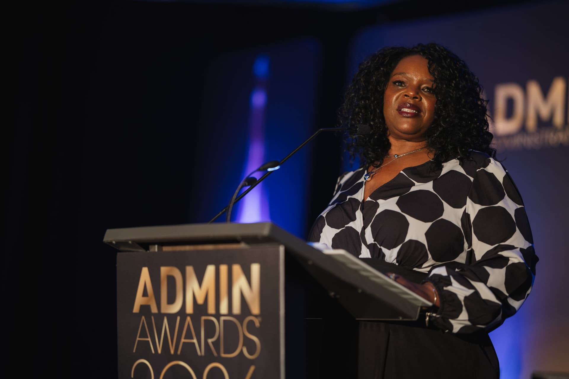 Speaker in a polka-dot blouse speaking passionately from the podium at Admin Awards 2026 at SFO Marriott Waterfront, Burlingame by JY Studio