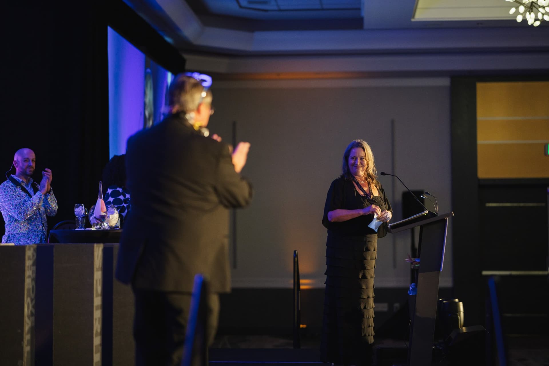 Guest with a microphone addressing an honoree on stage from the audience at Admin Awards 2026 at SFO Marriott Waterfront, Burlingame by JY Studio