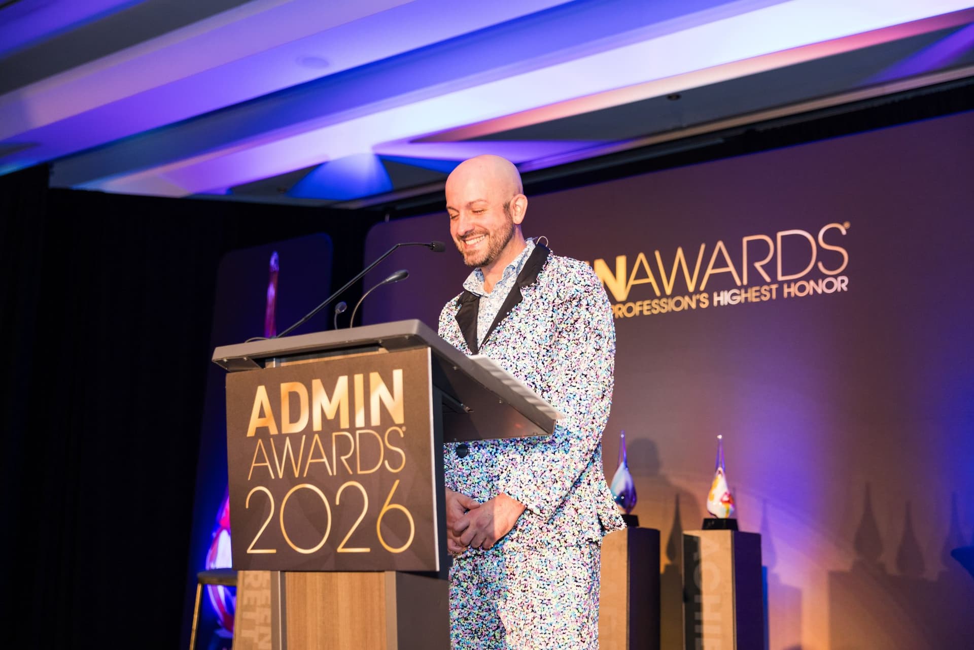 Emcee in a sequined suit smiling at the Admin Awards 2026 podium at Admin Awards 2026 at SFO Marriott Waterfront, Burlingame by JY Studio