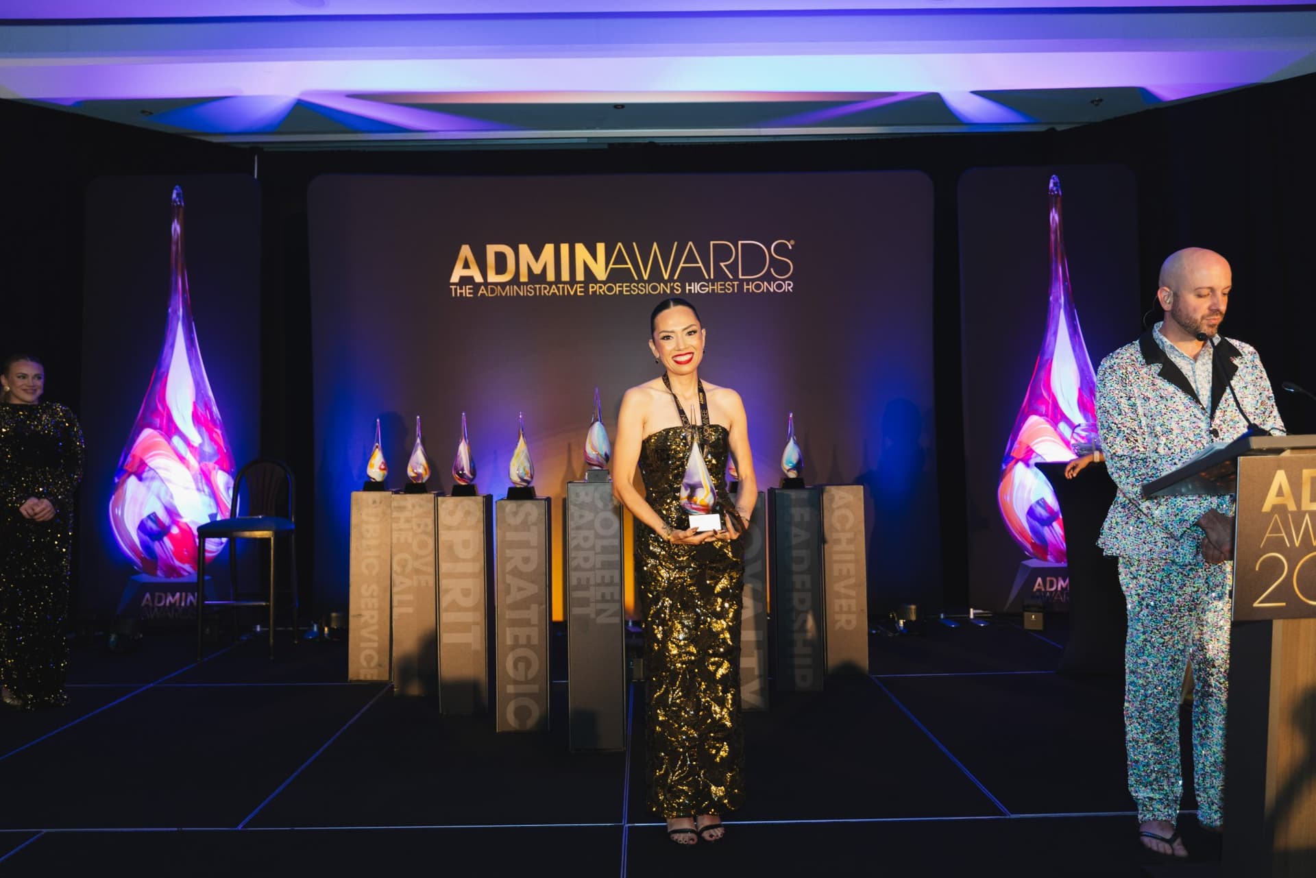 Award winner holding her trophy on stage beside the emcee at Admin Awards 2026 at SFO Marriott Waterfront, Burlingame by JY Studio