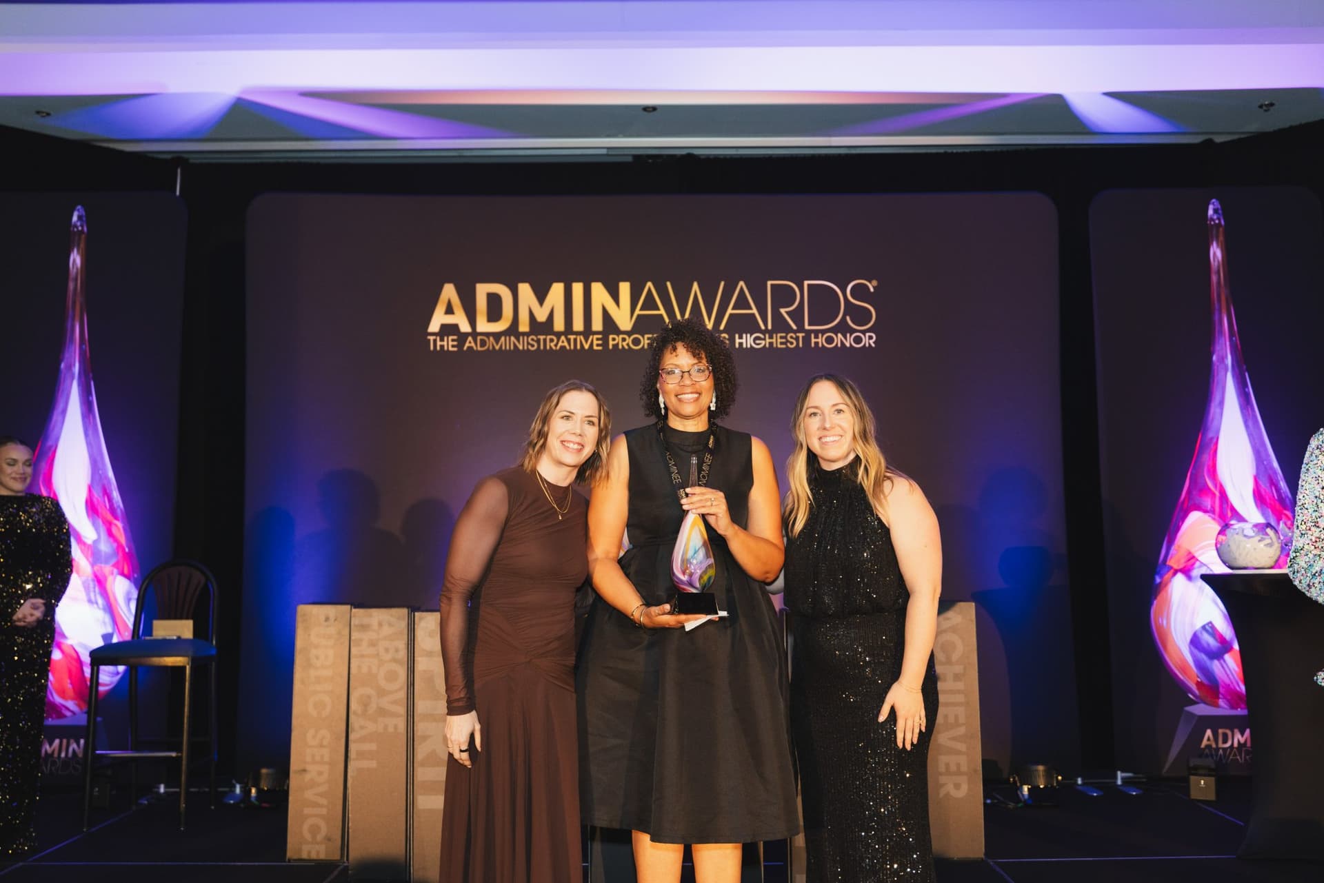 Award winner holding her trophy between two presenters on stage at Admin Awards 2026 at SFO Marriott Waterfront, Burlingame by JY Studio