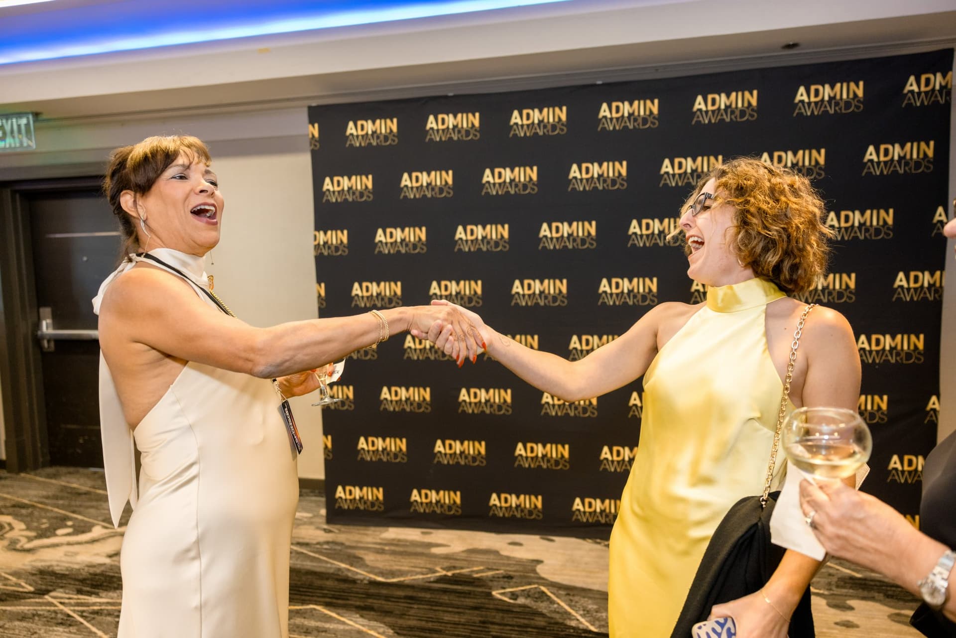Two guests laughing and shaking hands near the step-and-repeat backdrop at Admin Awards 2026 at SFO Marriott Waterfront, Burlingame by JY Studio