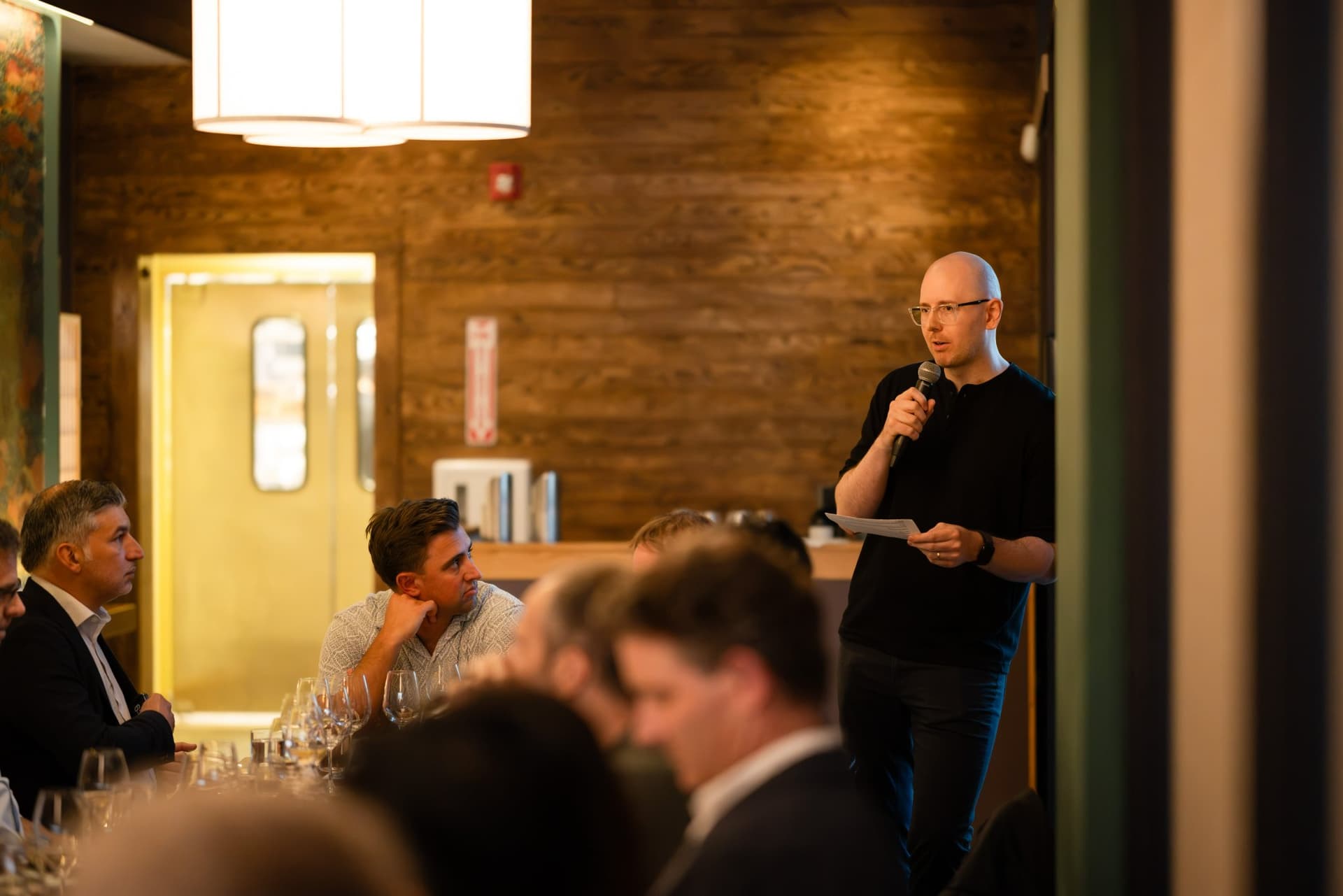 AI Insider host standing with a microphone to ask the table a question at the Business Insider AI Insider executive dinner The Great Coding Reset during SF Tech Week 2026 in San Francisco by JY Studio
