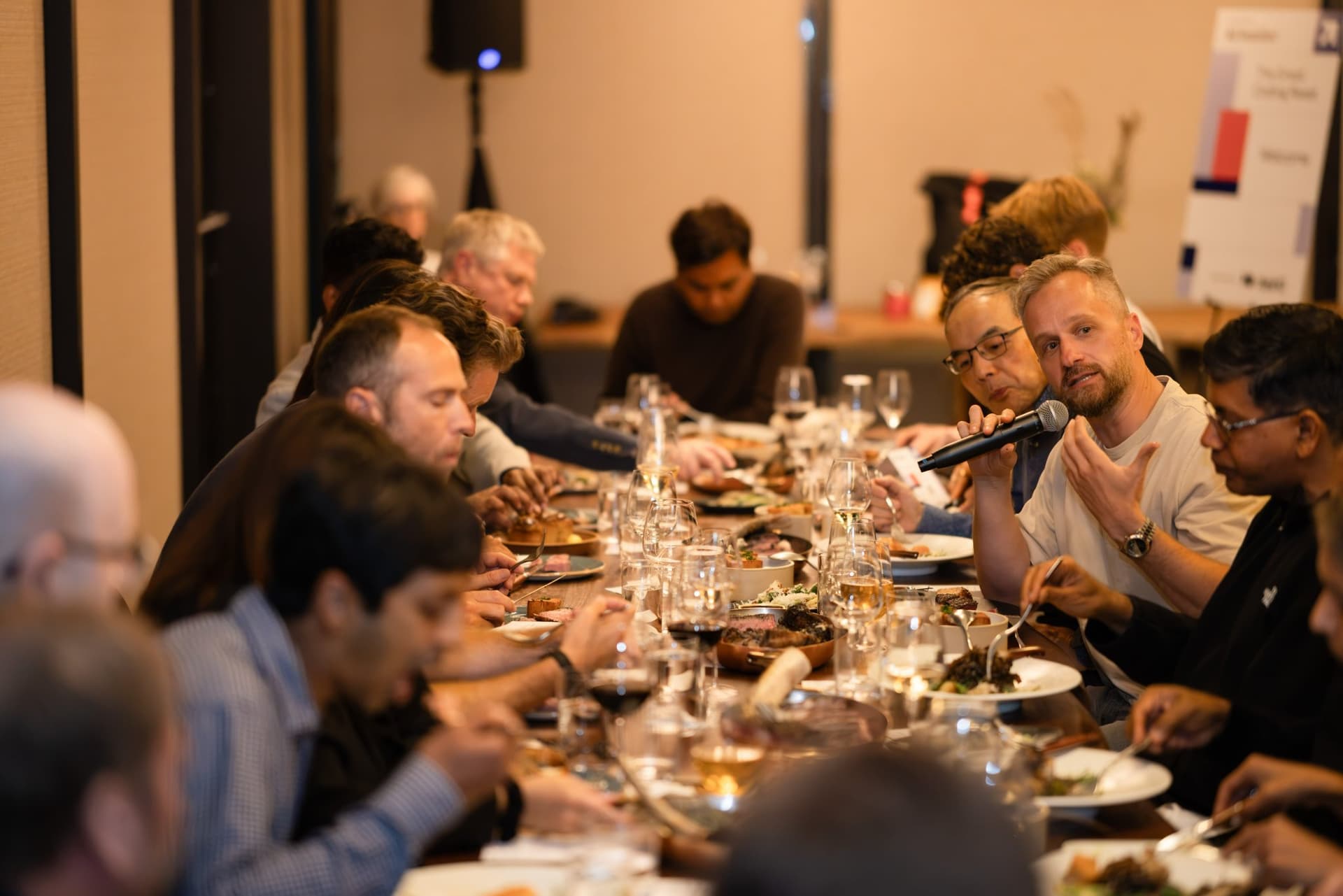 Executive speaking with a microphone over plates of food at the Business Insider AI Insider executive dinner The Great Coding Reset during SF Tech Week 2026 in San Francisco by JY Studio