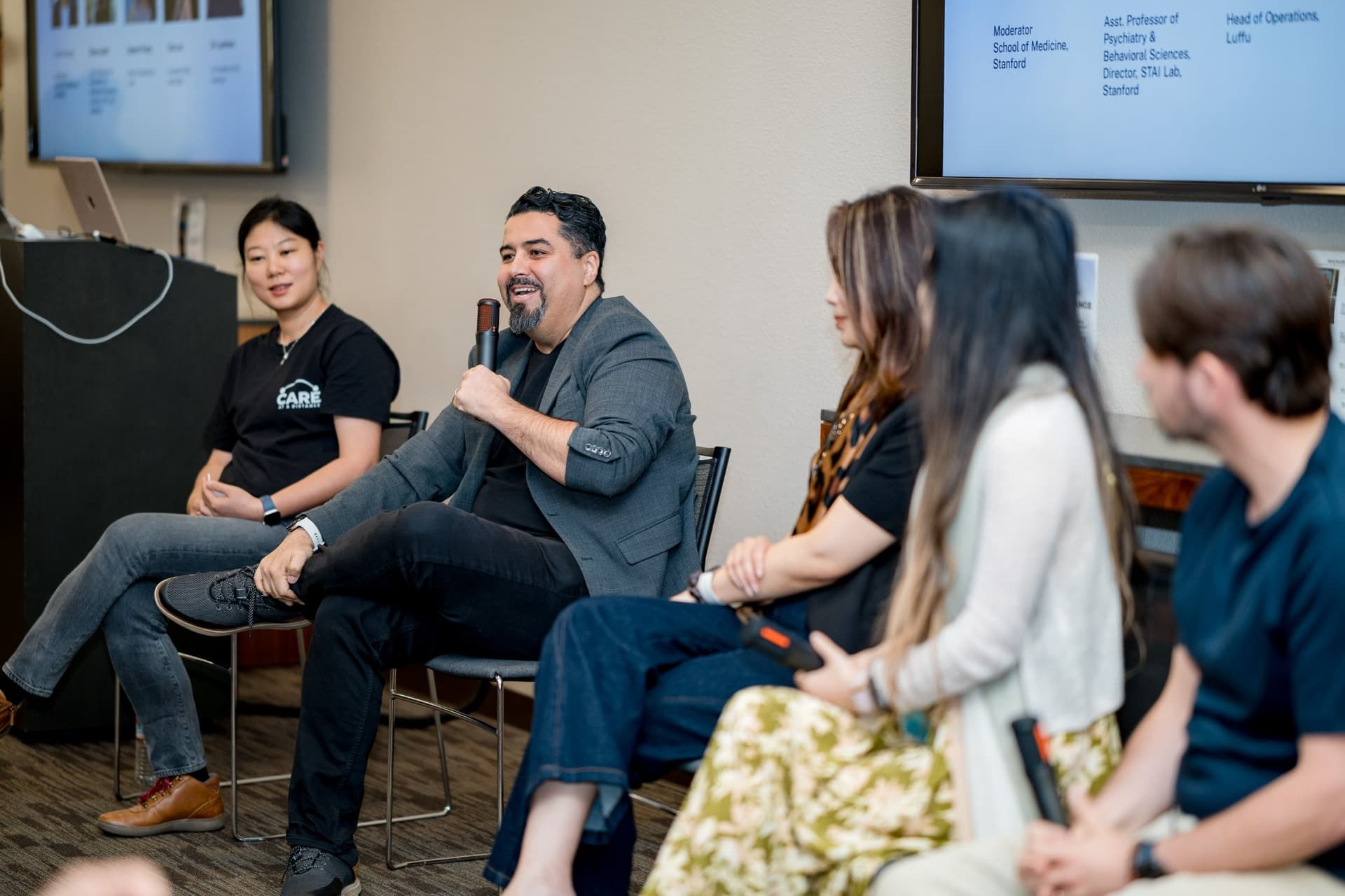 Panelist in a gray blazer speaking with a microphone during Panel 1 "Sensing Wellbeing from a Distance" at Care at a Distance: AI, Sensors, Robotics and the Future of Aging, SF Tech Week 2026 at EVGR, Stanford University by JY Studio