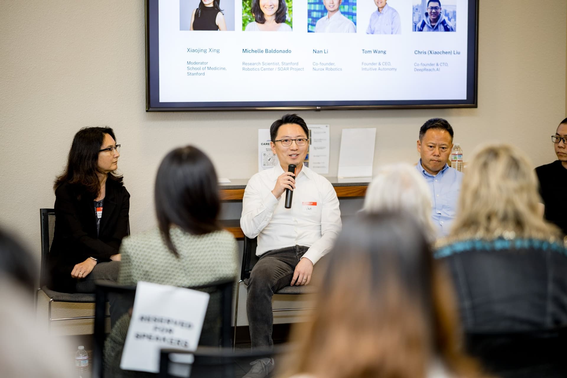 Panelist in a white shirt speaking during Panel 2 "Robotics for Independent Living" with the robotics panel slide behind at Care at a Distance: AI, Sensors, Robotics and the Future of Aging, SF Tech Week 2026 at EVGR, Stanford University by JY Studio