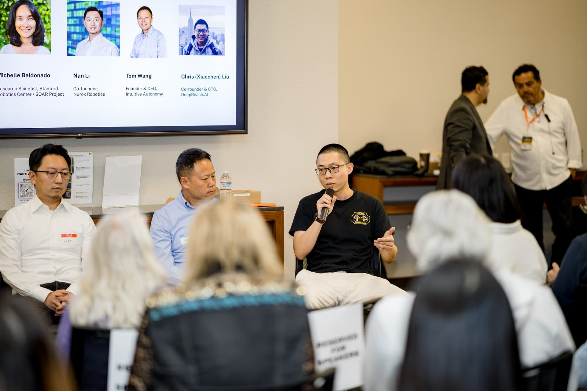 Panelist in a black T-shirt speaking about home robotics during Panel 2 "Robotics for Independent Living" at Care at a Distance: AI, Sensors, Robotics and the Future of Aging, SF Tech Week 2026 at EVGR, Stanford University by JY Studio