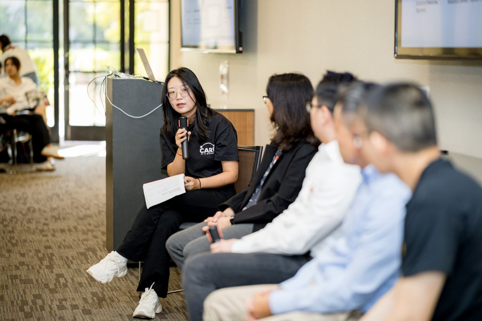 Moderator in a Care at a Distance T-shirt asking a question to the Panel 2 "Robotics for Independent Living" panelists at Care at a Distance: AI, Sensors, Robotics and the Future of Aging, SF Tech Week 2026 at EVGR, Stanford University by JY Studio
