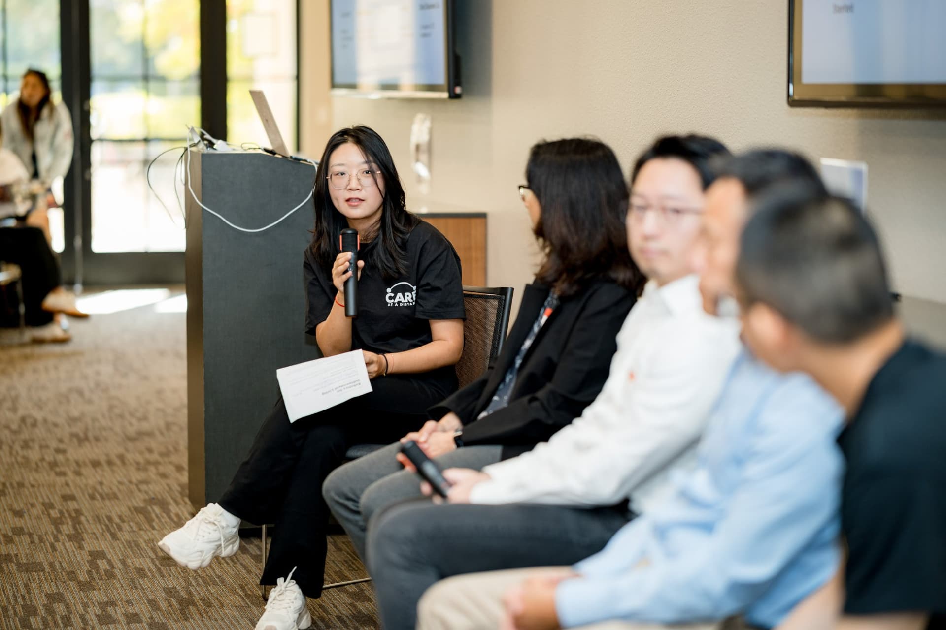 Moderator reading from a question card during Panel 2 "Robotics for Independent Living" at Care at a Distance: AI, Sensors, Robotics and the Future of Aging, SF Tech Week 2026 at EVGR, Stanford University by JY Studio