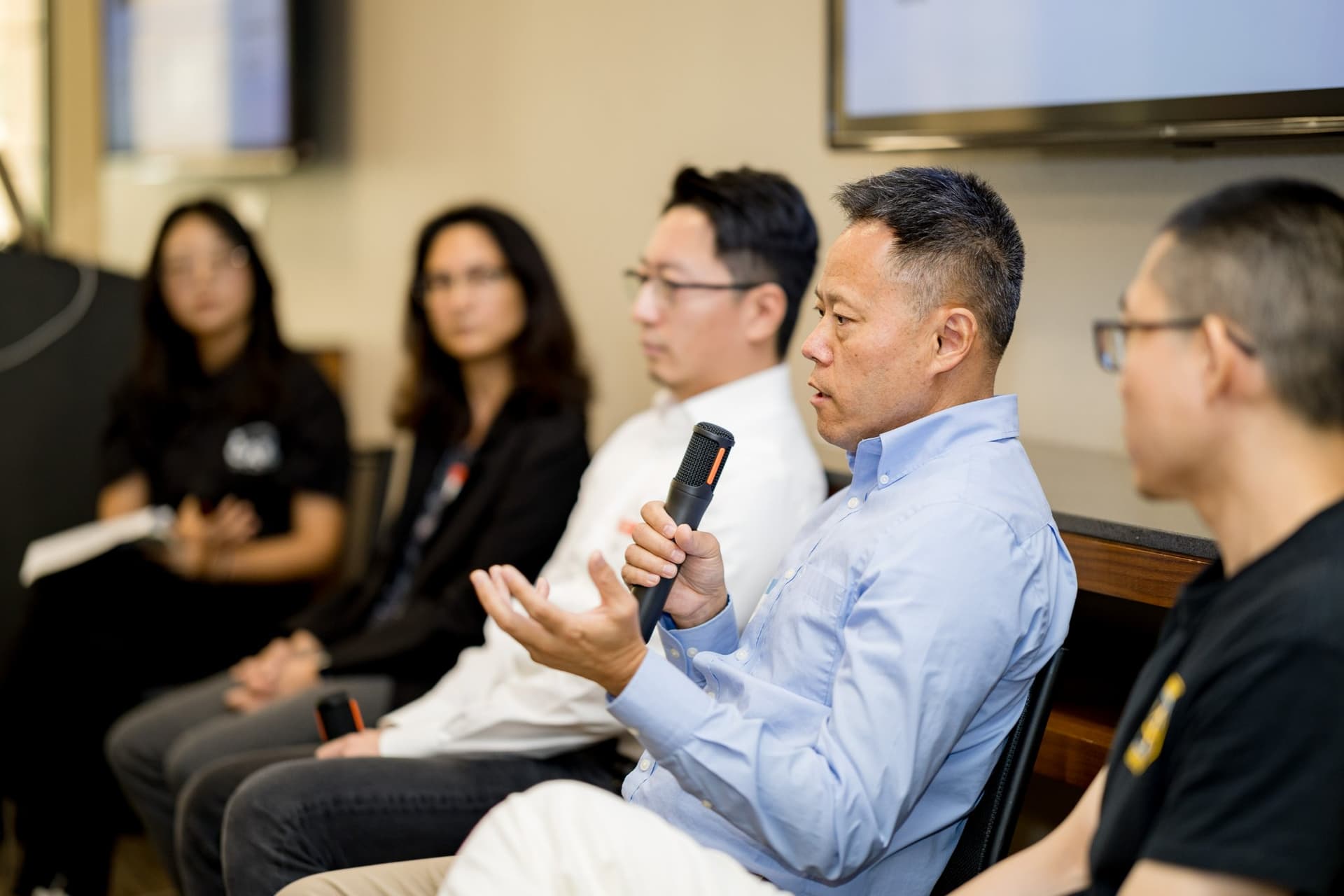 Panelist in a light blue shirt gesturing with a microphone during Panel 2 "Robotics for Independent Living" at Care at a Distance: AI, Sensors, Robotics and the Future of Aging, SF Tech Week 2026 at EVGR, Stanford University by JY Studio