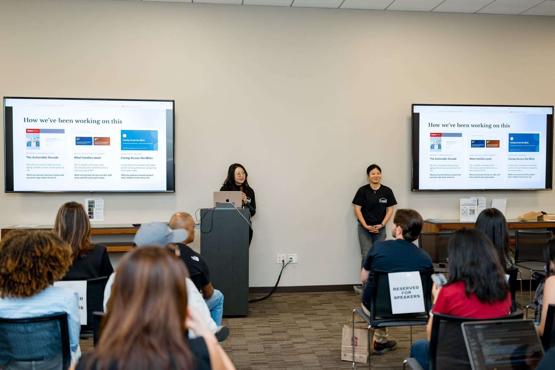 Event host presenting the Care at a Distance opening slides from the podium at Care at a Distance: AI, Sensors, Robotics and the Future of Aging, SF Tech Week 2026 at EVGR, Stanford University by JY Studio