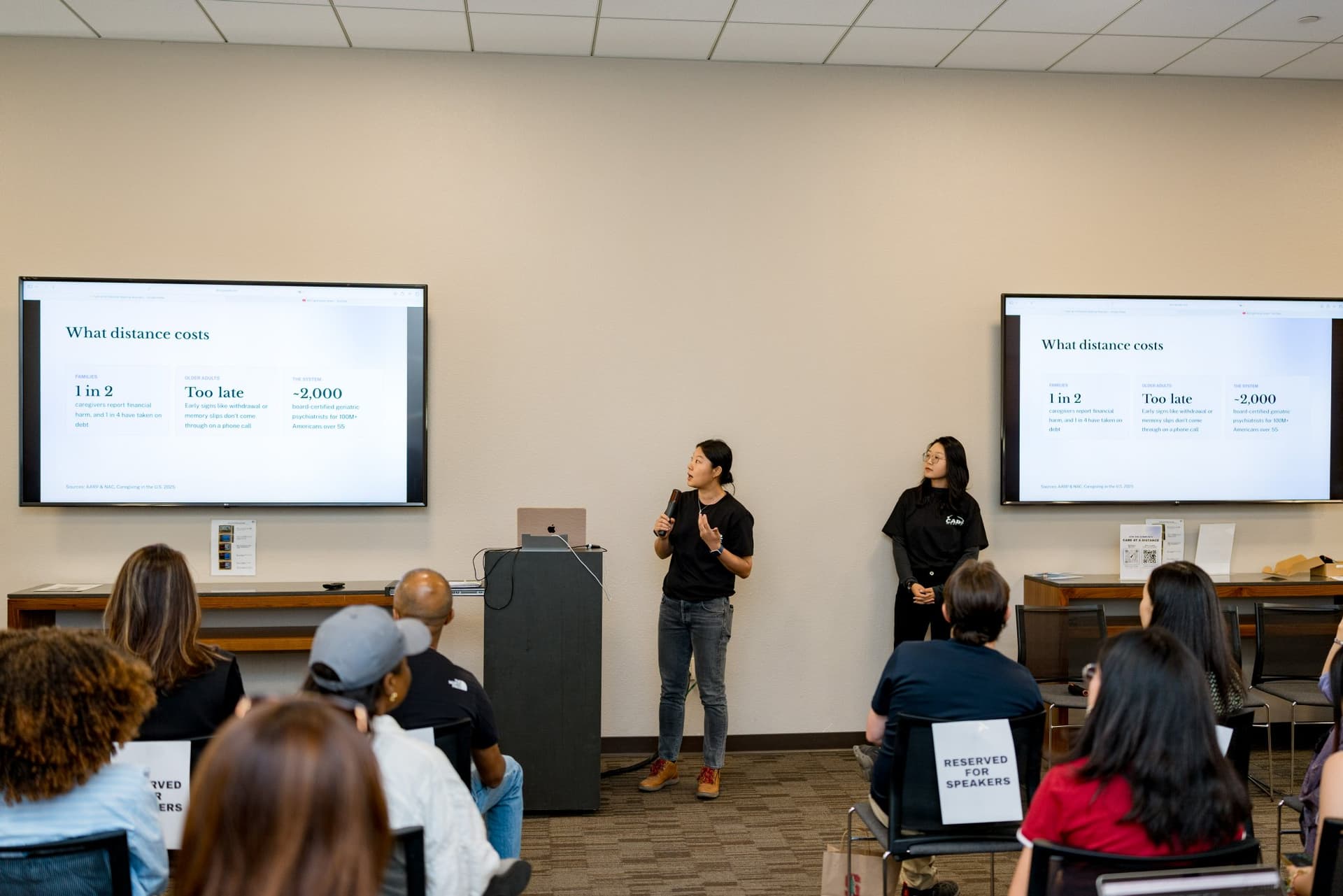 Hosts presenting the "What distance costs" slide on long-distance caregiving at Care at a Distance: AI, Sensors, Robotics and the Future of Aging, SF Tech Week 2026 at EVGR, Stanford University by JY Studio
