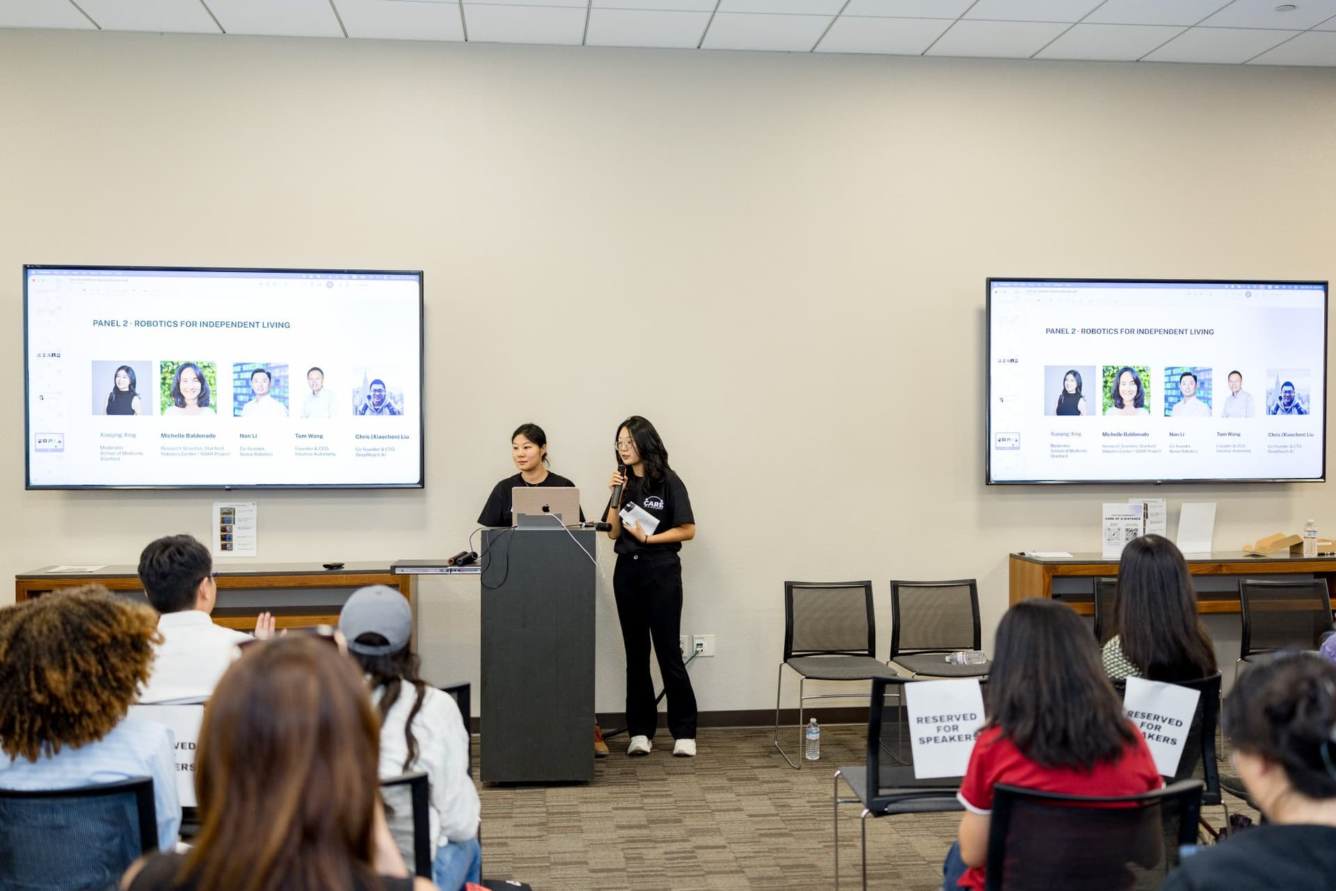 Hosts introducing Panel 2 "Robotics for Independent Living" at the podium at Care at a Distance: AI, Sensors, Robotics and the Future of Aging, SF Tech Week 2026 at EVGR, Stanford University by JY Studio