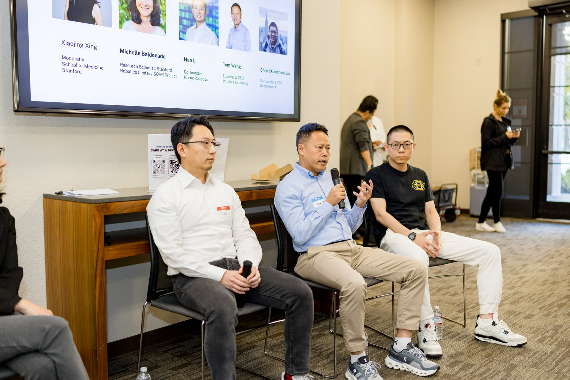 Three panelists of Panel 2 "Robotics for Independent Living" seated in front of the speaker slide at Care at a Distance: AI, Sensors, Robotics and the Future of Aging, SF Tech Week 2026 at EVGR, Stanford University by JY Studio