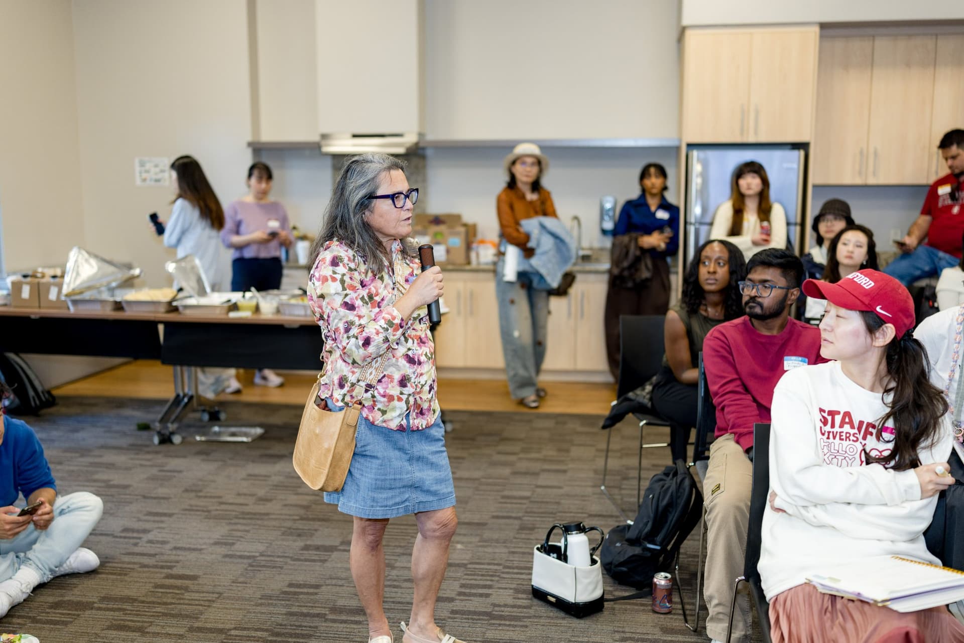 Attendee in a floral blouse asking the robotics panel a question with a microphone at Care at a Distance: AI, Sensors, Robotics and the Future of Aging, SF Tech Week 2026 at EVGR, Stanford University by JY Studio