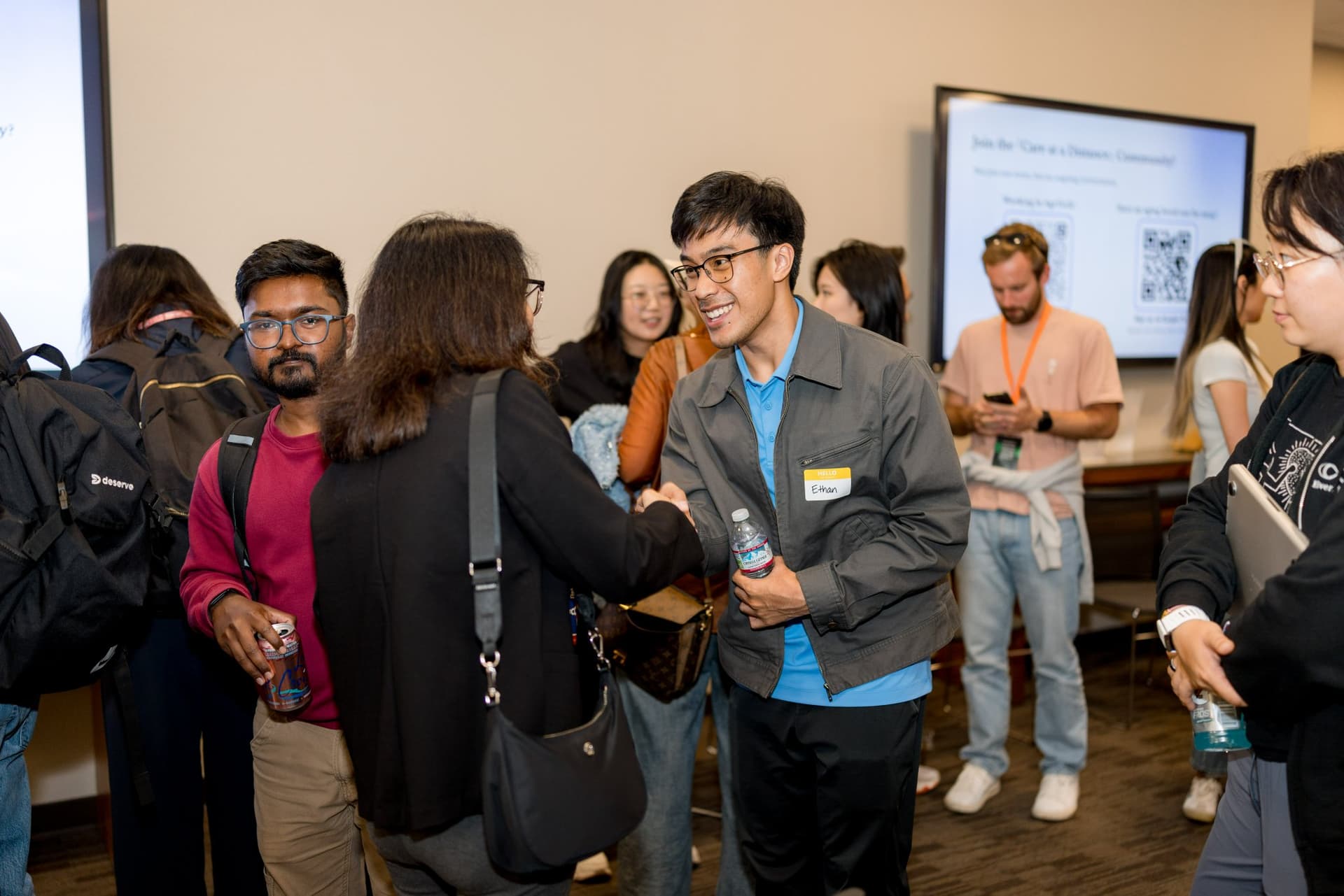 Founders shaking hands during networking beside the Join the Care at a Distance Community slide at Care at a Distance: AI, Sensors, Robotics and the Future of Aging, SF Tech Week 2026 at EVGR, Stanford University by JY Studio
