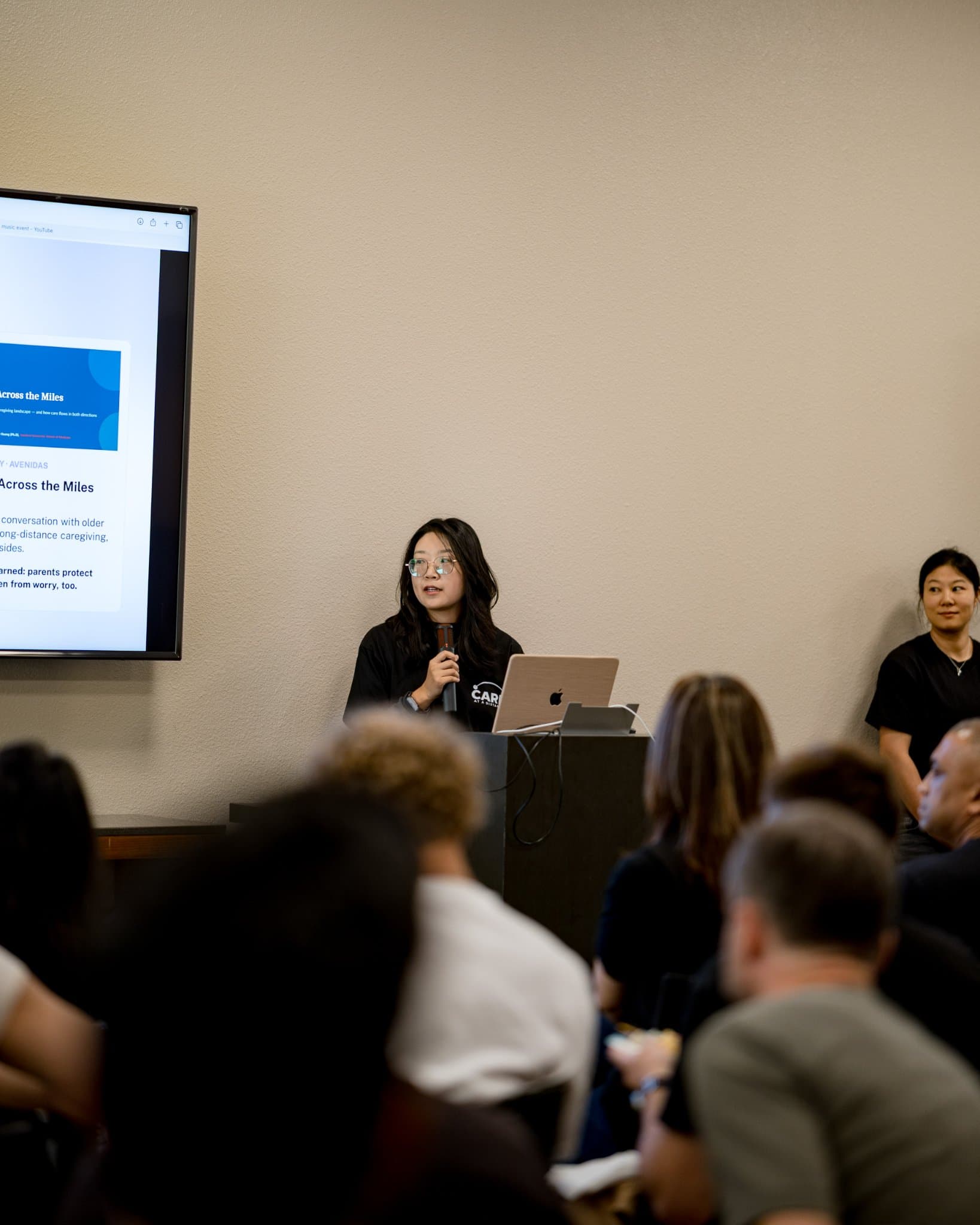 Vertical shot of a host speaking at the podium at Care at a Distance: AI, Sensors, Robotics and the Future of Aging, SF Tech Week 2026 at EVGR, Stanford University by JY Studio